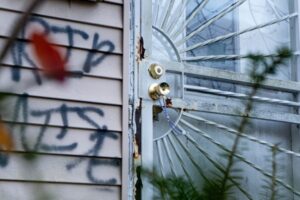 A rosary hangs on the doorknob of the home that once belonged to Alicia Fernnadze, with “RIP Wise” spray-painted on the exterior in memory of her late fiancé. Photo credit: Cydni Elledge/Outlier Media