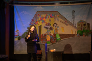 Director Karilu Alarcon Forshee gives the curtain speech at the beginning of the performance in front of the backdrop painted by Elton Monroy Dura