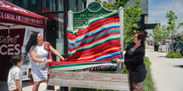 Two women unveiling a green Michigan Historic Site marker reading "Dra. Lucile Gajec" by pulling away a red, green, and blue striped serape, with a young boy watching beside them.