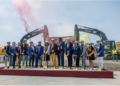 Group of about fourteen people in hard hats holding ceremonial gold shovels at a groundbreaking event, with excavators and colored smoke in the background