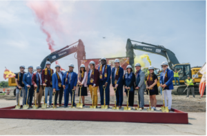 Group of about fourteen people in hard hats holding ceremonial gold shovels at a groundbreaking event, with excavators and colored smoke in the background