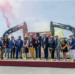 Group of about fourteen people in hard hats holding ceremonial gold shovels at a groundbreaking event, with excavators and colored smoke in the background
