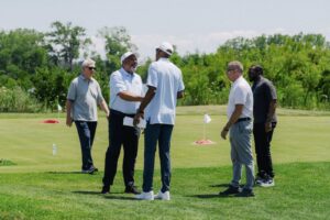 Ed Siaje, president of Bank of America Detroit, greets participants on a golf course during an outdoor event. Five men stand together on the green grass with trees and golf markers visible in the background
