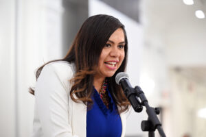 Close-up of a woman in a blue top and white blazer smiling and speaking into a microphone. 