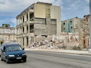 A pile of rubble sits beside a partially demolished multi-story building on a Havana street, with weathered pastel buildings and a blue car passing in the foreground.