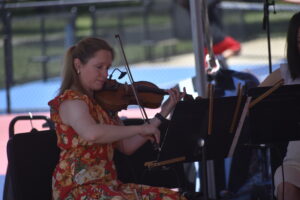 Close-up of a woman playing violin outdoors in a red and yellow floral dress, reading sheet music on a stand.