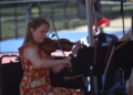Close-up of a woman playing violin outdoors in a red and yellow floral dress, reading sheet music on a stand.