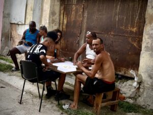 A group of men playing dominoes at an outdoor table against a rusted metal door in Havana, with onlookers seated nearby against a weathered wall.