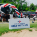 The entrance to Bank of America's Golf With Us event features a large red, white, and blue balloon arch above the event sign, with a white shuttle bus and event staff greeting young participants arriving for the program
