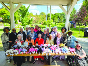 Large group of about 20 women posing together under a park pavilion, with rows of dressed dolls lined up on a table in front of them, greenery and trees visible in the background.