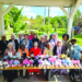 Large group of about 20 women posing together under a park pavilion, with rows of dressed dolls lined up on a table in front of them, greenery and trees visible in the background.