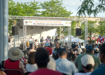 Wide view of a crowd watching a band perform on an outdoor stage under a banner reading "We Tell These Truths: All Humans Are Created Equal," trees and buildings in the background.