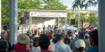 Wide view of a crowd watching a band perform on an outdoor stage under a banner reading "We Tell These Truths: All Humans Are Created Equal," trees and buildings in the background.
