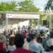 Wide view of a crowd watching a band perform on an outdoor stage under a banner reading "We Tell These Truths: All Humans Are Created Equal," trees and buildings in the background.