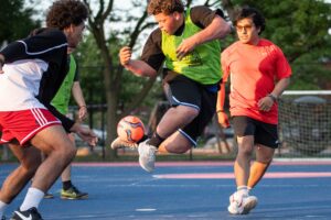 Three young players vie for the soccer ball during intense active play, with spectators and field fencing visible in the background