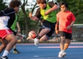 Three young players vie for the soccer ball during intense active play, with spectators and field fencing visible in the background