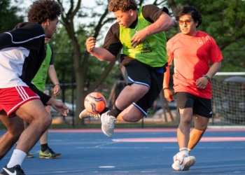 Three young players vie for the soccer ball during intense active play, with spectators and field fencing visible in the background