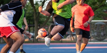 Three young players vie for the soccer ball during intense active play, with spectators and field fencing visible in the background