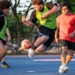 Three young players vie for the soccer ball during intense active play, with spectators and field fencing visible in the background