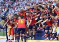 Spain's national team celebrating on a podium platform with medals around their necks, with two men in suits standing near the stage under a "World Cup 2026" banner.