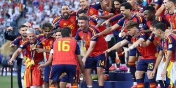Spain's national team celebrating on a podium platform with medals around their necks, with two men in suits standing near the stage under a "World Cup 2026" banner.