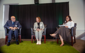 Three panelists seated in chairs on a small stage with artificial turf flooring, one speaking into a microphone while holding papers, name tags reading "Osvaldo Rivera," "Michelle Farkkonen," and "Stephanie Peña" visible.