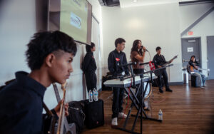 Teen band performing indoors at an event, with a singer, guitarists, and a keyboardist in matching black outfits with red heart patches, an "Alliance Catholic Credit Union" sponsor slide displayed on a screen behind them.