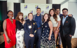 Group of eight people posing together and smiling in front of a "Southwest Detroit Business Association" step-and-repeat banner, including a woman in a police uniform.