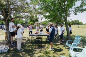 An instructor leads an outdoor group session under large shade trees, addressing a circle of participants wearing Golf With Us t-shirts gathered around a table, with a residential building and green space visible in the background