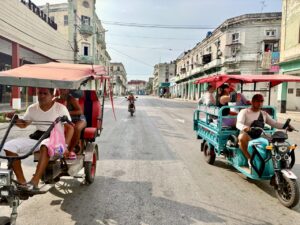 Motorized tricycle taxis carrying passengers pass each other on a wide Havana street lined with weathered colonial-style buildings.