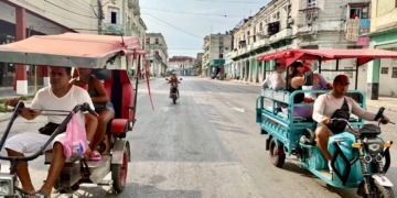 Motorized tricycle taxis carrying passengers pass each other on a wide Havana street lined with weathered colonial-style buildings.