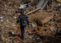 Emergency responder wearing a helmet and backpack walking across a large field of debris and rubble.