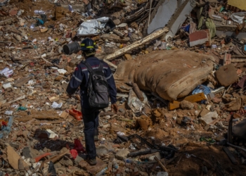 Emergency responder wearing a helmet and backpack walking across a large field of debris and rubble.
