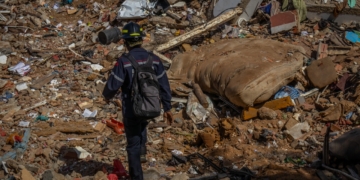 Emergency responder wearing a helmet and backpack walking across a large field of debris and rubble.