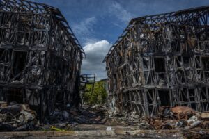 Two burned-out building skeletons reduced to charred metal framing, with wrecked vehicles in the foreground under a blue sky.