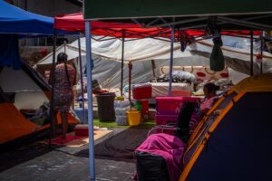 Makeshift living space under tarps and pop-up canopies, with a mattress, water containers, and personal belongings arranged on the ground, two people visible inside.