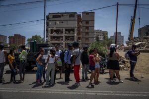 Crowd of people standing in a street watching a damaged, partially collapsed apartment building, with a crane and rubble visible in the background.