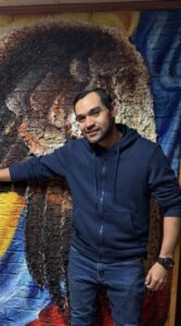 Man in a navy zip-up hoodie standing and smiling in front of a large textured mural featuring warm earth tones and blue accents.
