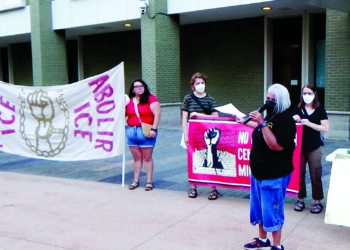 People standing outside a building holding banners reading "Abolish ICE" and "Abolir ICE," featuring an illustration of a raised fist breaking chains; some hold protest signs and one person speaks into a microphone