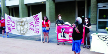 People standing outside a building holding banners reading "Abolish ICE" and "Abolir ICE," featuring an illustration of a raised fist breaking chains; some hold protest signs and one person speaks into a microphone