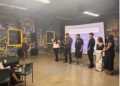 Multiple panelists stand in discussion in front of a projection screen reading "THREE WAYS TO TAKE ACTION" at an event space with distinctive yellow-framed windows and decorative skull murals on the walls