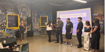Multiple panelists stand in discussion in front of a projection screen reading "THREE WAYS TO TAKE ACTION" at an event space with distinctive yellow-framed windows and decorative skull murals on the walls