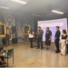 Multiple panelists stand in discussion in front of a projection screen reading "THREE WAYS TO TAKE ACTION" at an event space with distinctive yellow-framed windows and decorative skull murals on the walls