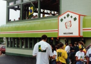 Exterior of a pub decorated with a Brazilian flag and a "Welcome" banner, with a crowd of soccer fans gathered outside and visible through an upstairs window.