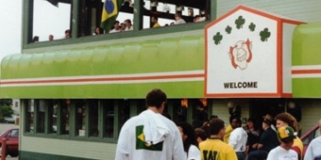 Exterior of a pub decorated with a Brazilian flag and a "Welcome" banner, with a crowd of soccer fans gathered outside and visible through an upstairs window.
