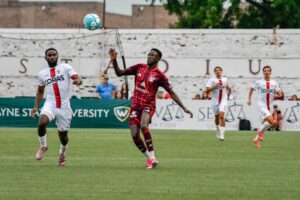 Live soccer action showing a DCFC player in maroon AlumniFct jersey competing with an opposing player in white CORAS jersey for a blue soccer ball, with additional players and spectators visible on the field