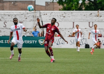 Live soccer action showing a DCFC player in maroon AlumniFct jersey competing with an opposing player in white CORAS jersey for a blue soccer ball, with additional players and spectators visible on the field