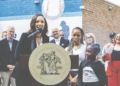 A woman speaks into a microphone at a large golden seal podium reading "Mayor of the City" at an outdoor event. Behind her stand school officials and students, with the baseball-themed mural visible on the brick building backdrop. Several officials and a young student in a California Athletic League shirt stand nearby