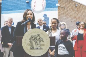 A woman speaks into a microphone at a large golden seal podium reading "Mayor of the City" at an outdoor event. Behind her stand school officials and students, with the baseball-themed mural visible on the brick building backdrop. Several officials and a young student in a California Athletic League shirt stand nearby