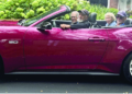 Four people ride together in a burgundy convertible classic car with white-wall tires, driving through a tree-lined residential street
